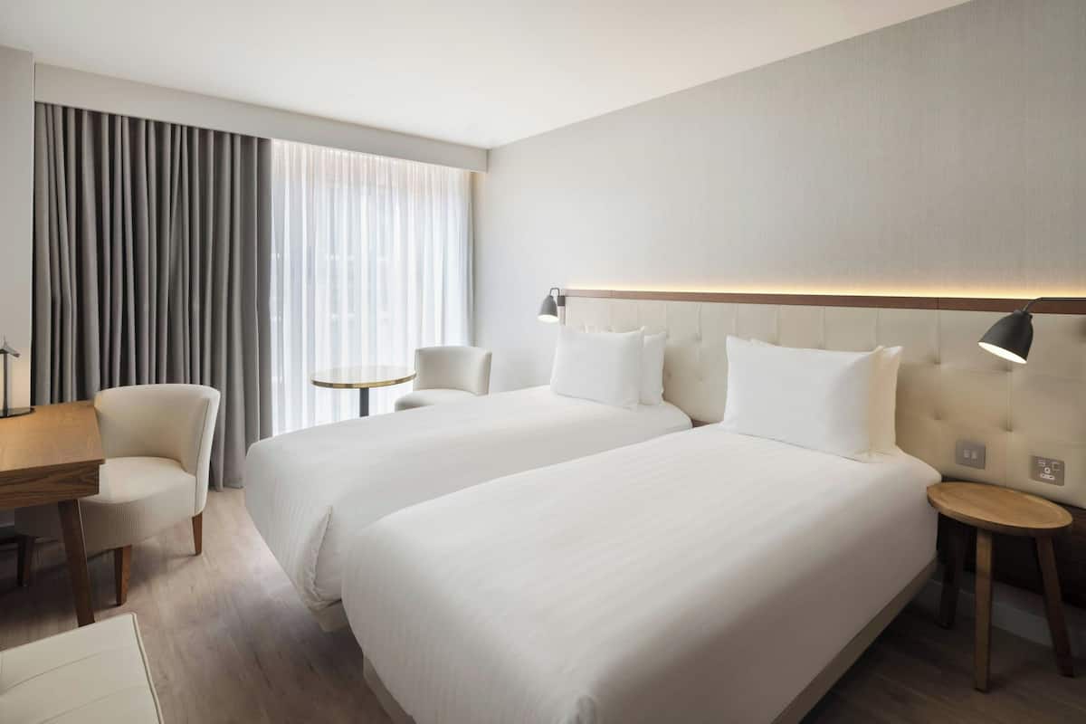 Ein Hotelzimmer mit zwei Betten, einem Schreibtisch, einem Stuhl, einer Lampe und Blick zum Fenster hinaus.