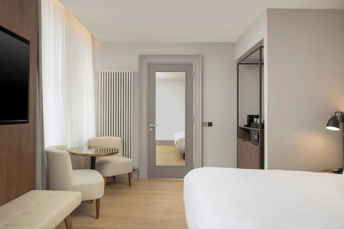 Ein Hotelzimmer mit einem Bett, einem runden Tisch mit zwei Stühlen, einem an der Wand montierten Fernseher, einem Spiegel und einer Tür zu einem weiteren Raum.