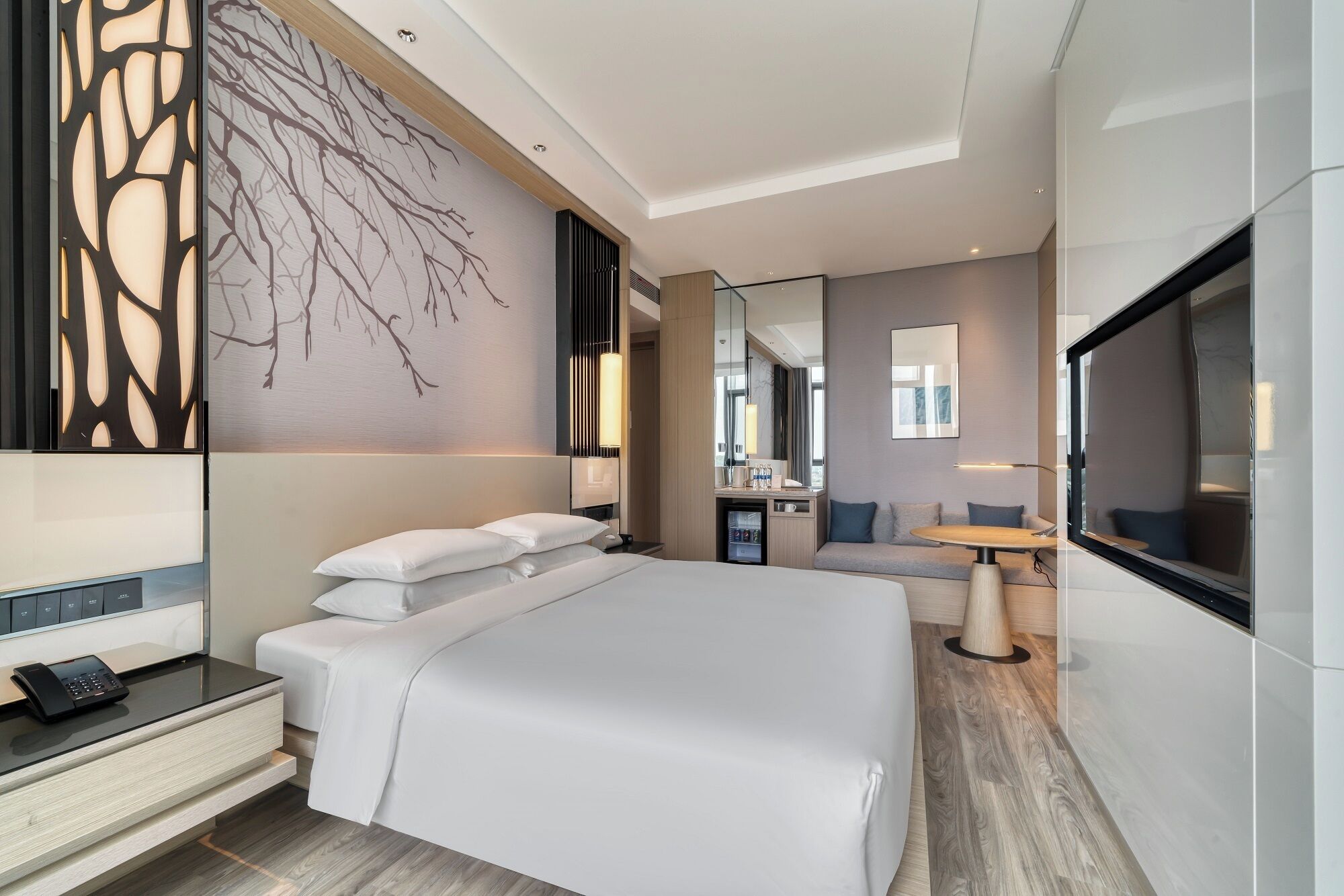 Quarto de hotel moderno com uma cama grande, uma escrivaninha com telefone, TV e uma área de estar com mesa e cadeiras.
