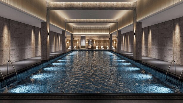 Une longue piscine rectangulaire intérieure, dotée d’un sol à motifs, entourée de murs et de colonnes en pierre, avec un escalier central et un espace détente.