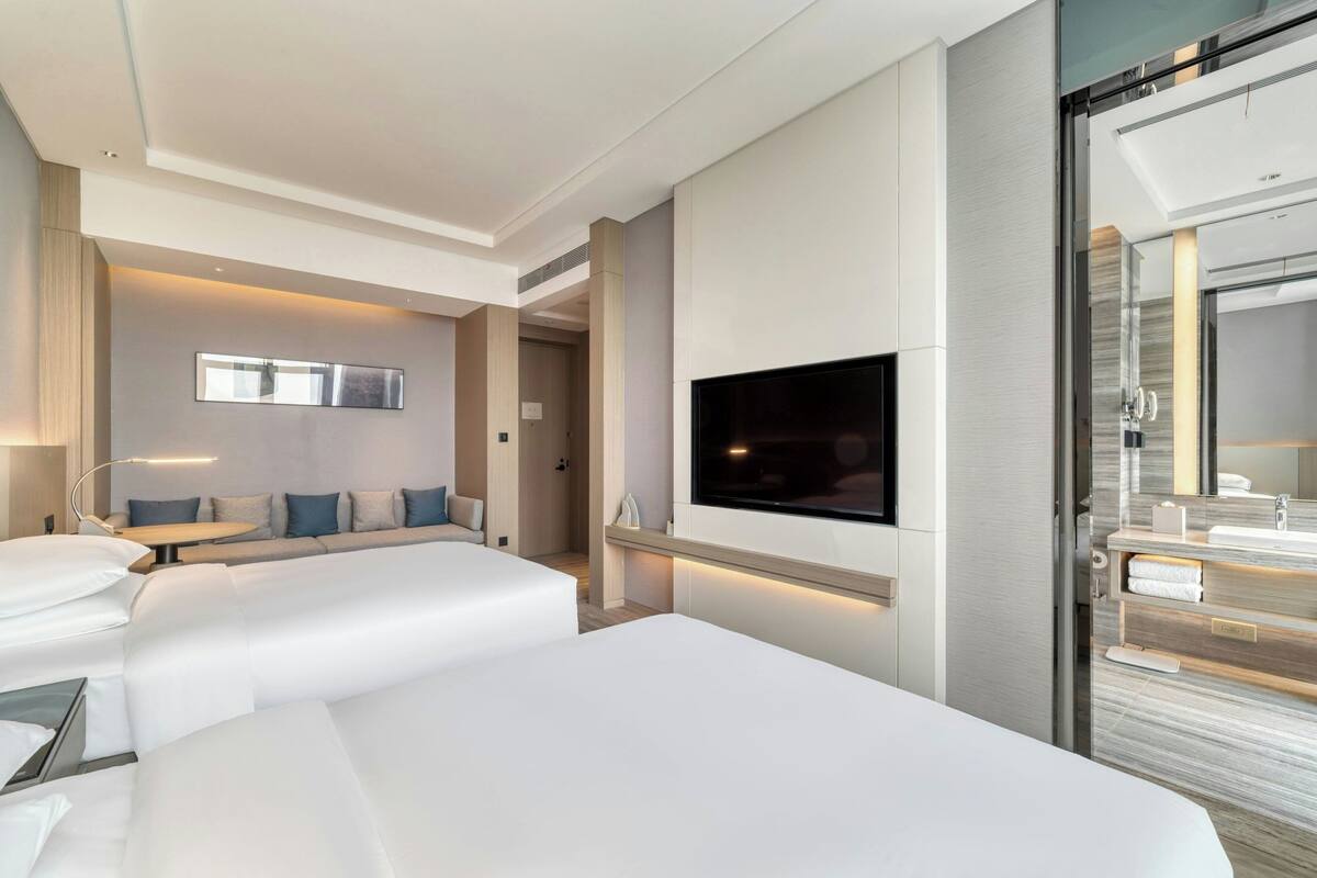 Una habitación de hotel moderna con dos camas, un televisor de pantalla plana, un baño con espejo y una zona de estar con sofá y cojines.