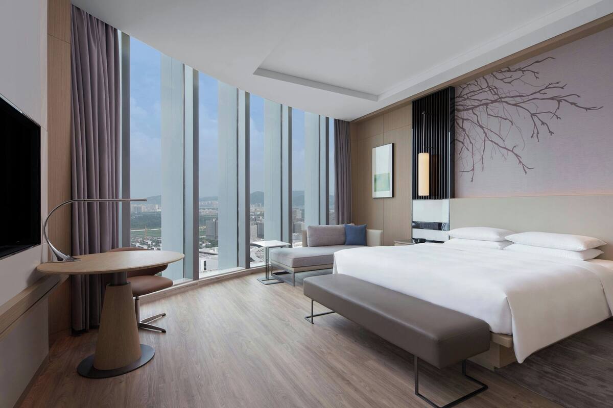 Habitación de hotel moderna con una cama grande, un escritorio, un banco y vistas de la ciudad a través de ventanales de piso a techo.