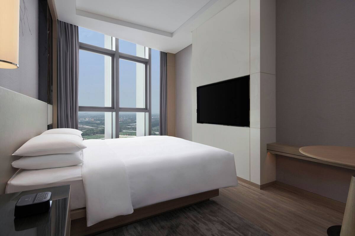 Una habitación de hotel moderna con una cama grande, un televisor de pantalla plana montado en la pared y vistas de la ciudad a través de un amplio ventanal.