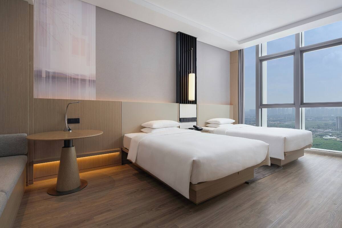 Habitación de hotel moderna con un ventanal grande, una mesa redonda, dos camas y una lámpara fijada a la pared.
