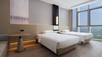 A modern hotel room with a large window, a round table, two beds, and a wall-mounted light fixture.