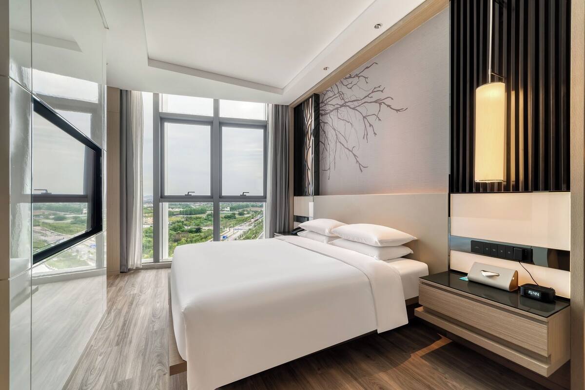 Una habitación de hotel moderna con una cama grande, una mesita de noche con lámpara y vistas a la ciudad a través de amplios ventanales.