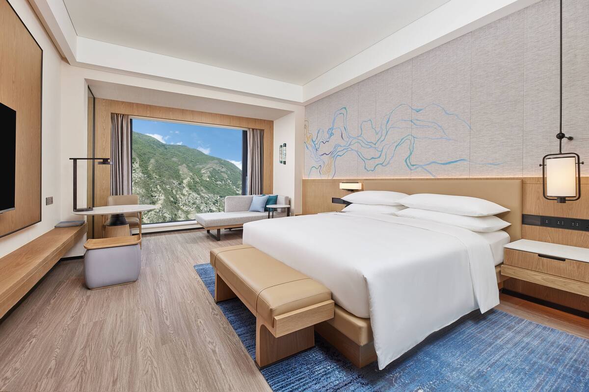 Habitación de hotel moderna con una cama grande, un escritorio, un banco y vistas a un paisaje montañoso.