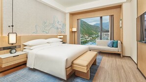 Une chambre d’hôtel avec un grand lit, une table de chevet avec une lampe, un banc, une télévision et une vue sur les montagnes.