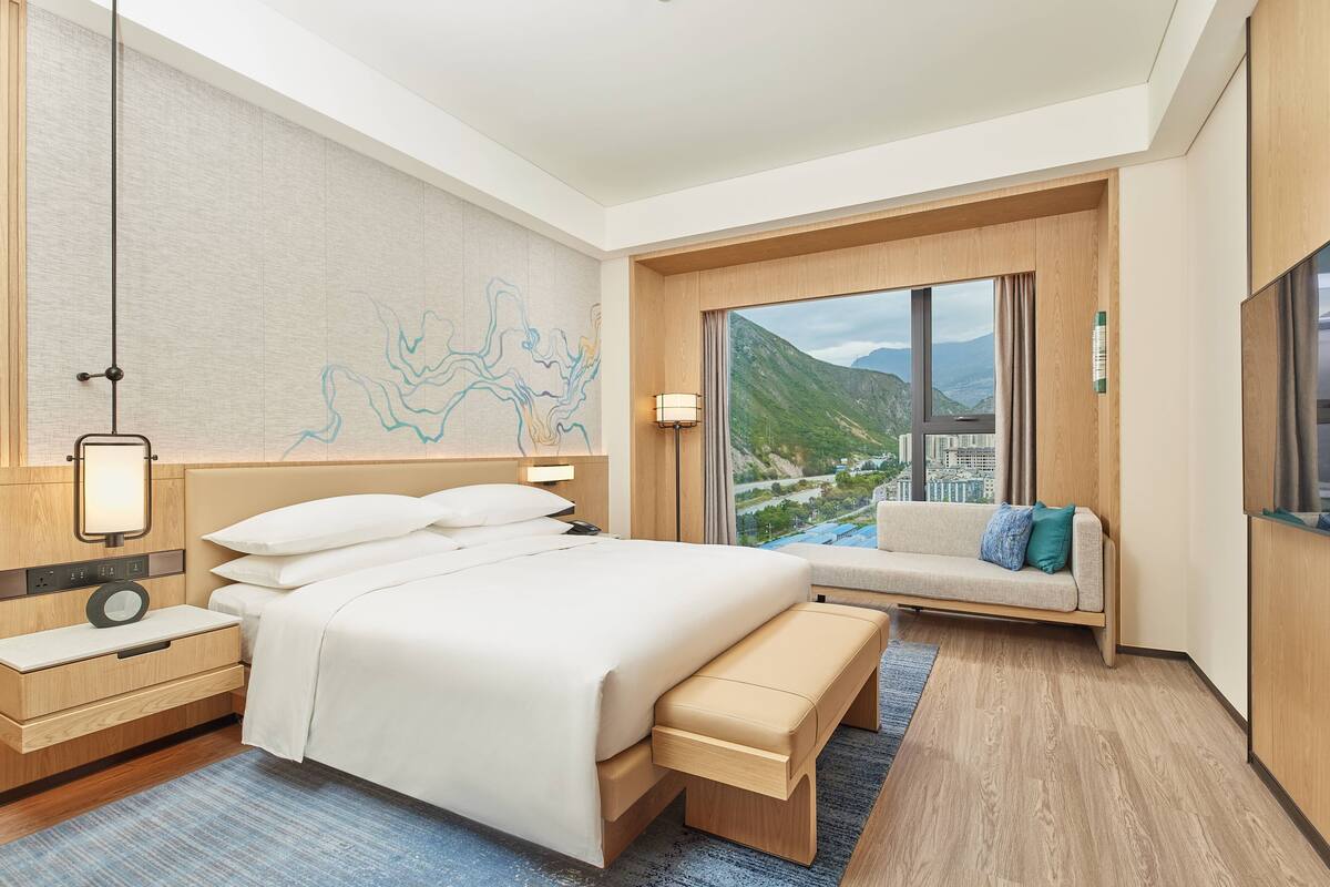 Una habitación de hotel con una cama grande, una mesita de noche con lámpara, un banco, un televisor y vistas a las montañas.