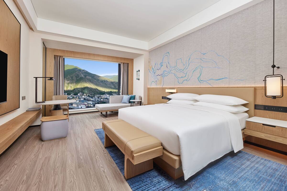 Una habitación de hotel moderna con una cama grande, un escritorio, un banco y vistas a un paisaje montañoso.