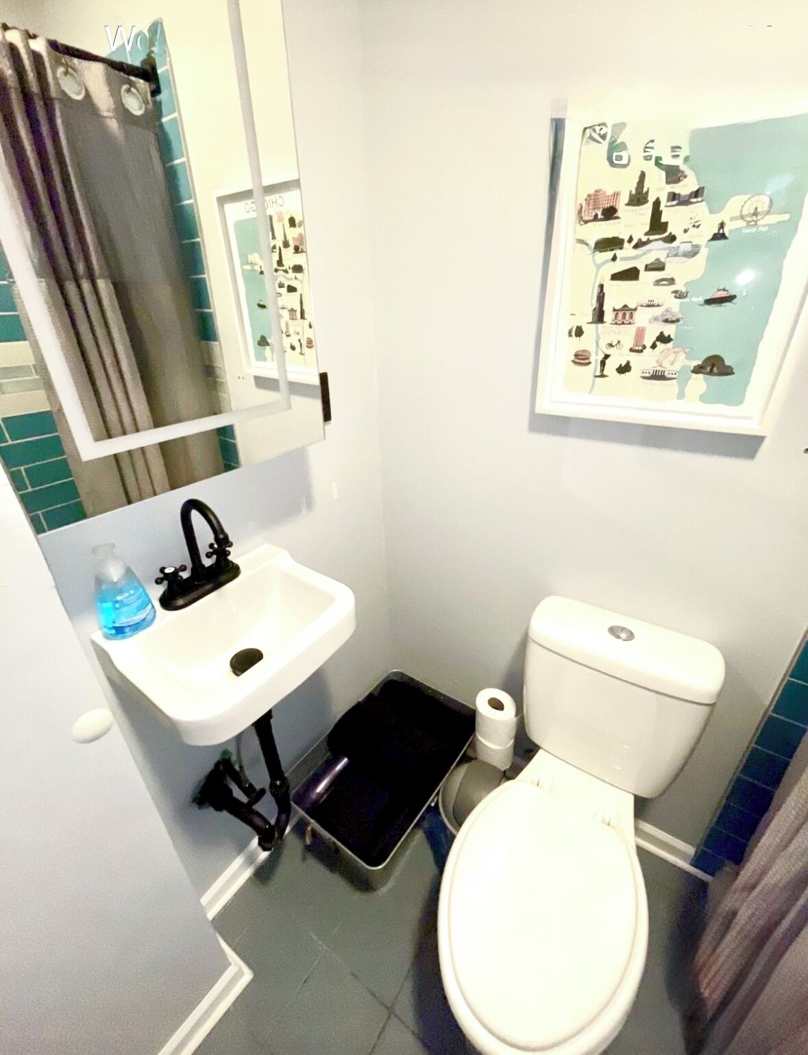 Une petite salle de bain avec un lavabo blanc, un robinet noir et des toilettes. On y trouve un rideau de douche, un miroir et un tableau encadré accroché au mur.