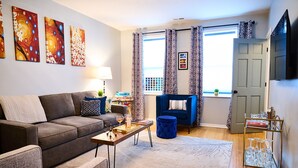 A living room with a sofa, coffee table, and a blue ottoman. There are abstract paintings on the wall, a window with curtains, and a door.