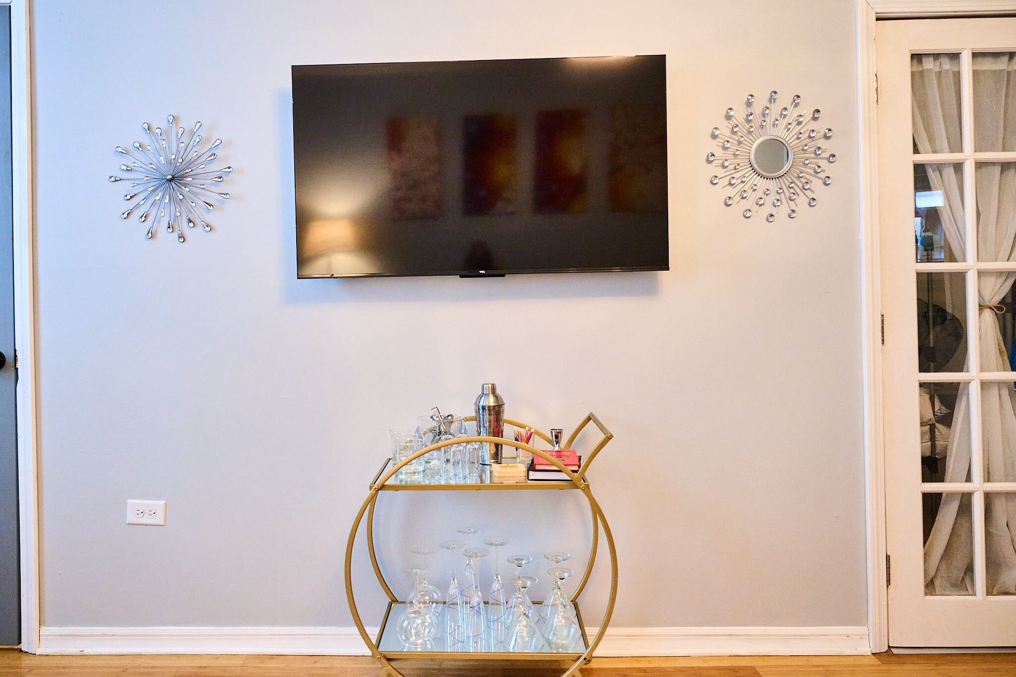 Quarto com televisão de tela plana na parede, carrinho de bar com tampo de vidro, copos e garrafas, e arte decorativa na parede.