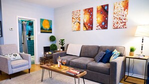 A living room with a grey sofa, a coffee table with books and a glass, a side table with a lamp, and a door leading to a bathroom.