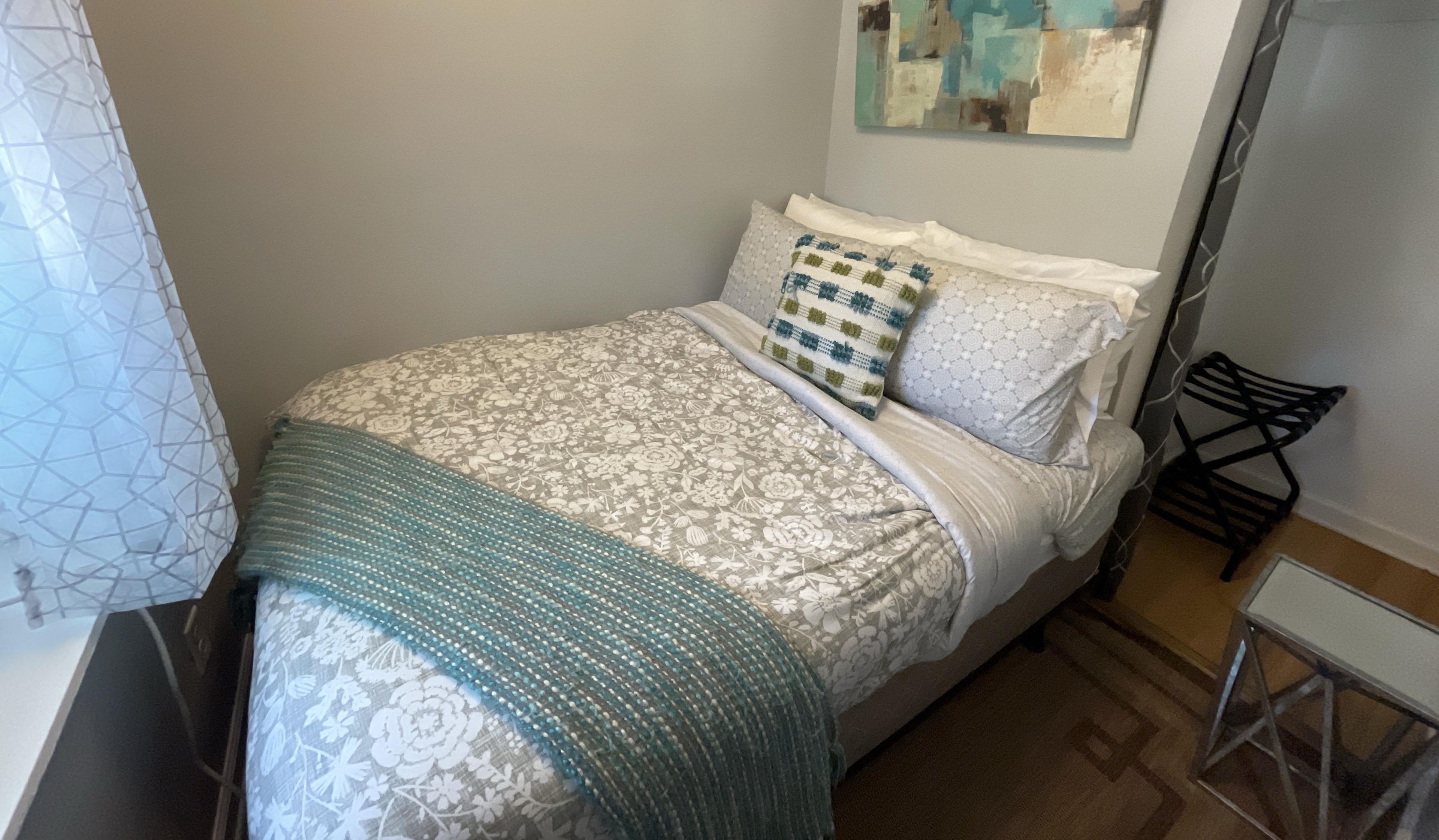 Un dormitorio con cama, almohadas, colcha, una silla y una mesita.