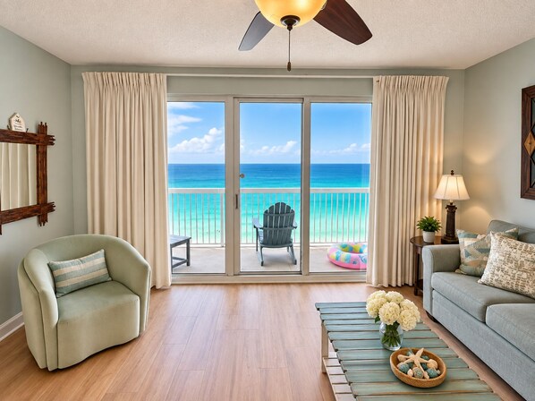 A room with a view of the ocean, a ceiling fan, a sofa, a chair, and a small table with flowers.
