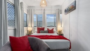 A hotel room with a bed, two bedside tables with lamps, a red and grey sofa, and large windows with curtains.