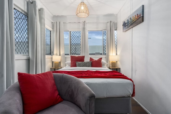 A hotel room with a bed, two bedside tables with lamps, a red and grey sofa, and large windows with curtains.