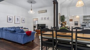 A modern living room with a blue sofa, dining table, and chairs. There is a TV, air conditioning unit, and framed pictures on the walls.