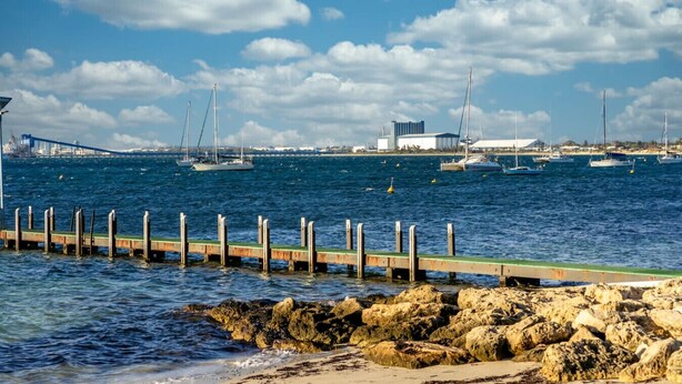 A coastal marina with boats, a bridge in the background, and a rocky shoreline.