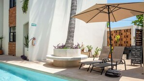 Een zwembadgedeelte met ligbedden, een plantenbak met een palmboom en een bord waarop de openingstijden en activiteiten bij het zwembad staan.