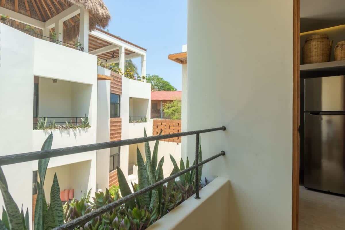 A modern building with multiple balconies, some with plants. There is a metal railing in the foreground, and a stainless steel refrigerator is visible in the background.