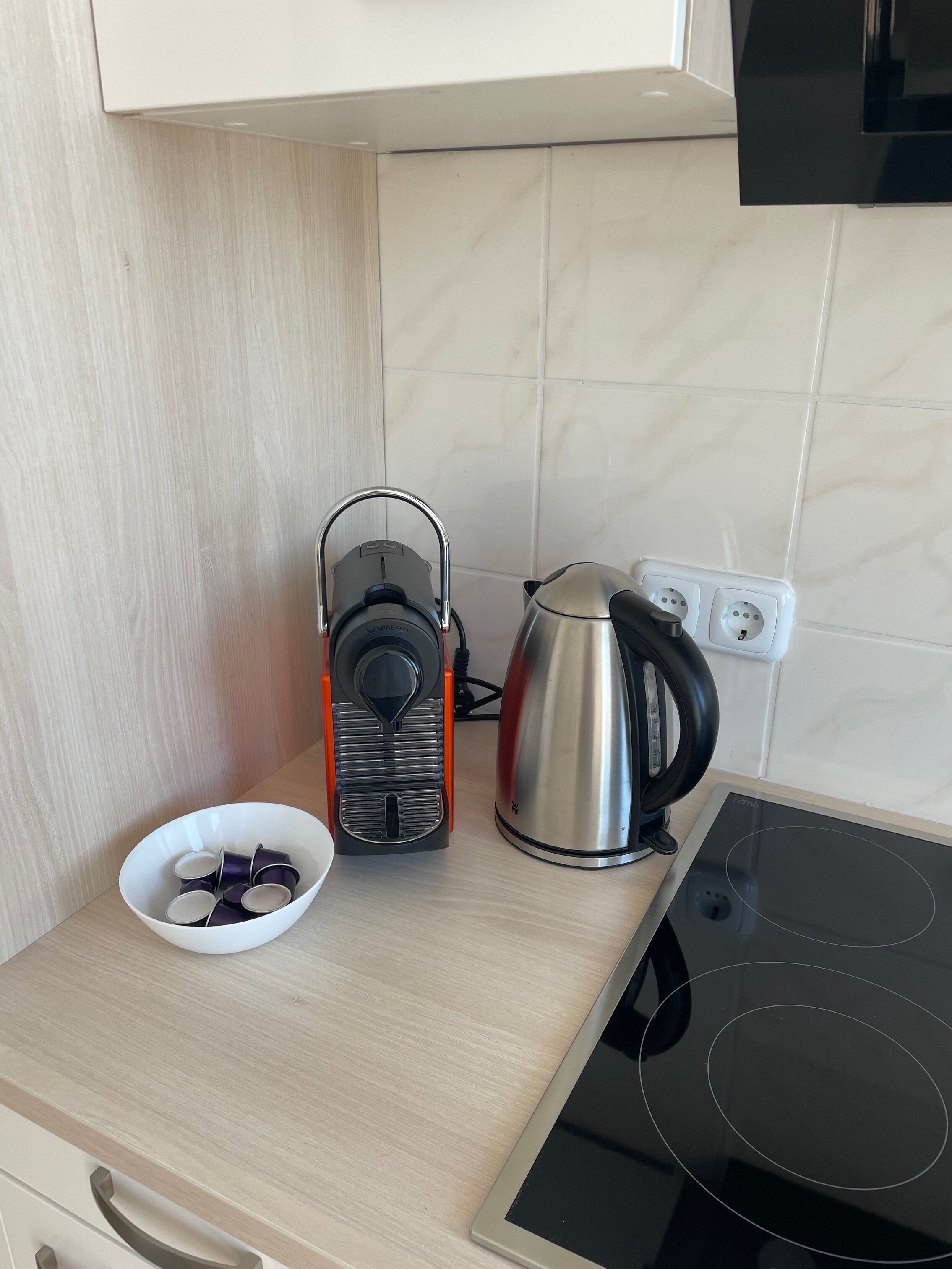 A kitchen with a kettle, a coffee maker, and a bowl of cups on a countertop.