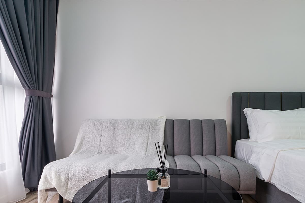A modern hotel room with a grey sofa, a small round table with a plant and a bottle, and a bed with white bedding.