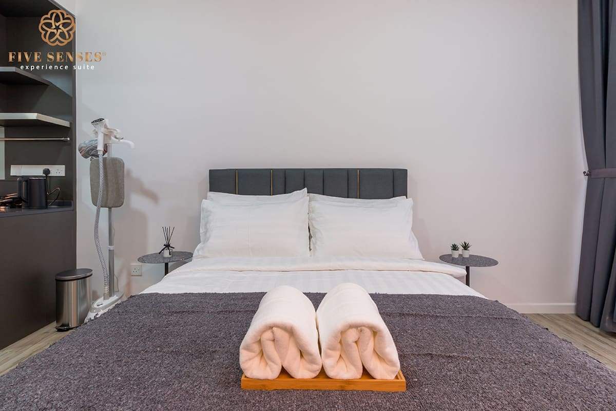 A neatly made bed with a grey headboard, a grey blanket, and two rolled towels on a wooden tray. There is a small table with a plant and a lamp on the side.