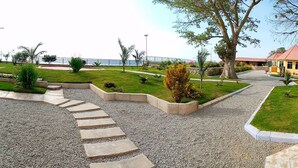 A landscaped garden with a gravel path, a yellow building, and a view of the sea. - SANDMARK HOTEL SERENGETI  (Bariadi)