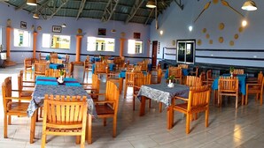 A spacious dining area with wooden tables and chairs, a high ceiling with metal beams, and large windows allowing natural light. - SANDMARK HOTEL SERENGETI  (Bariadi)