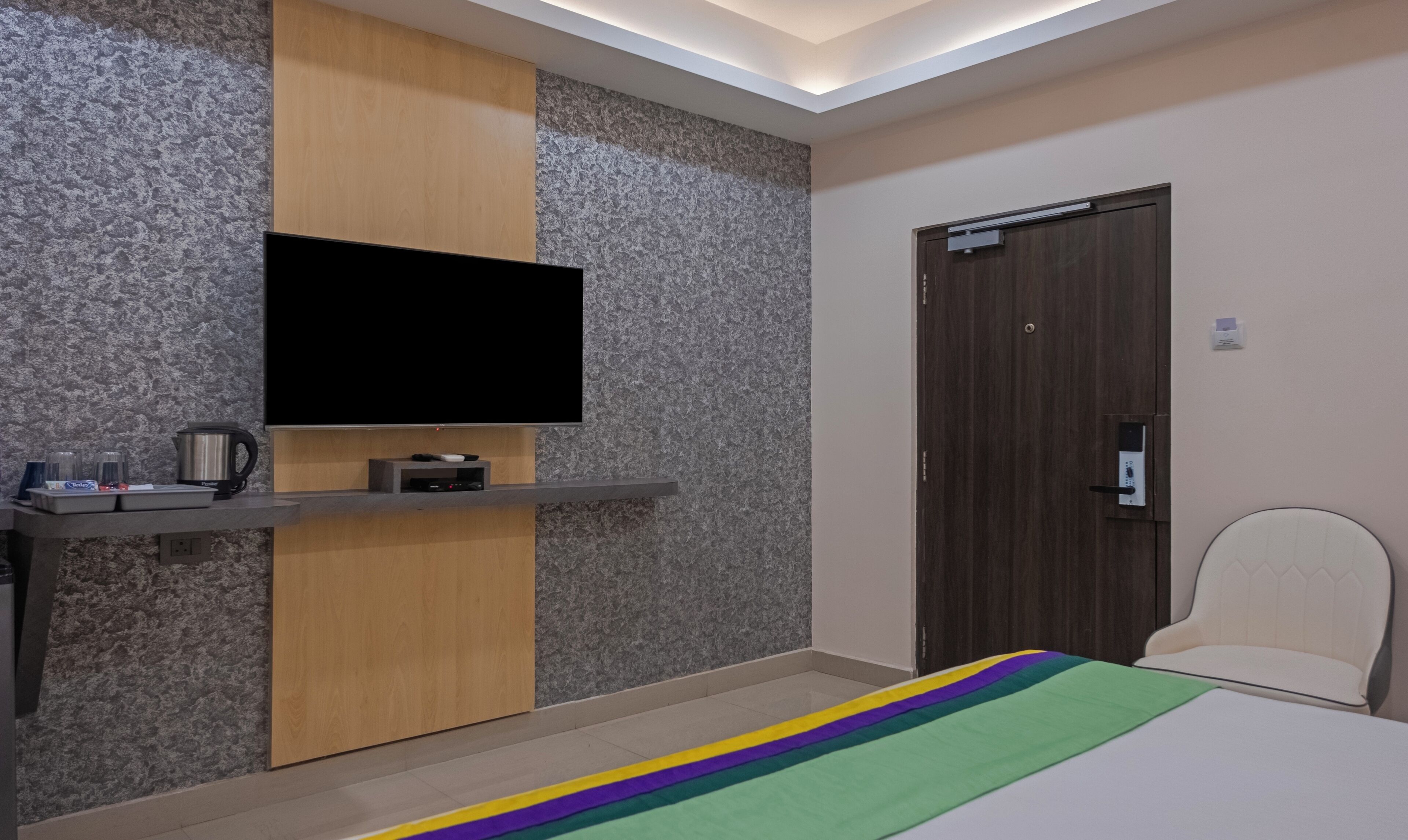 Habitación de hotel con televisión de pantalla plana montada en la pared, un mueble de madera debajo, una mesita con tetera y vasos, una cama con colcha a rayas de arcoíris y una puerta con manija.