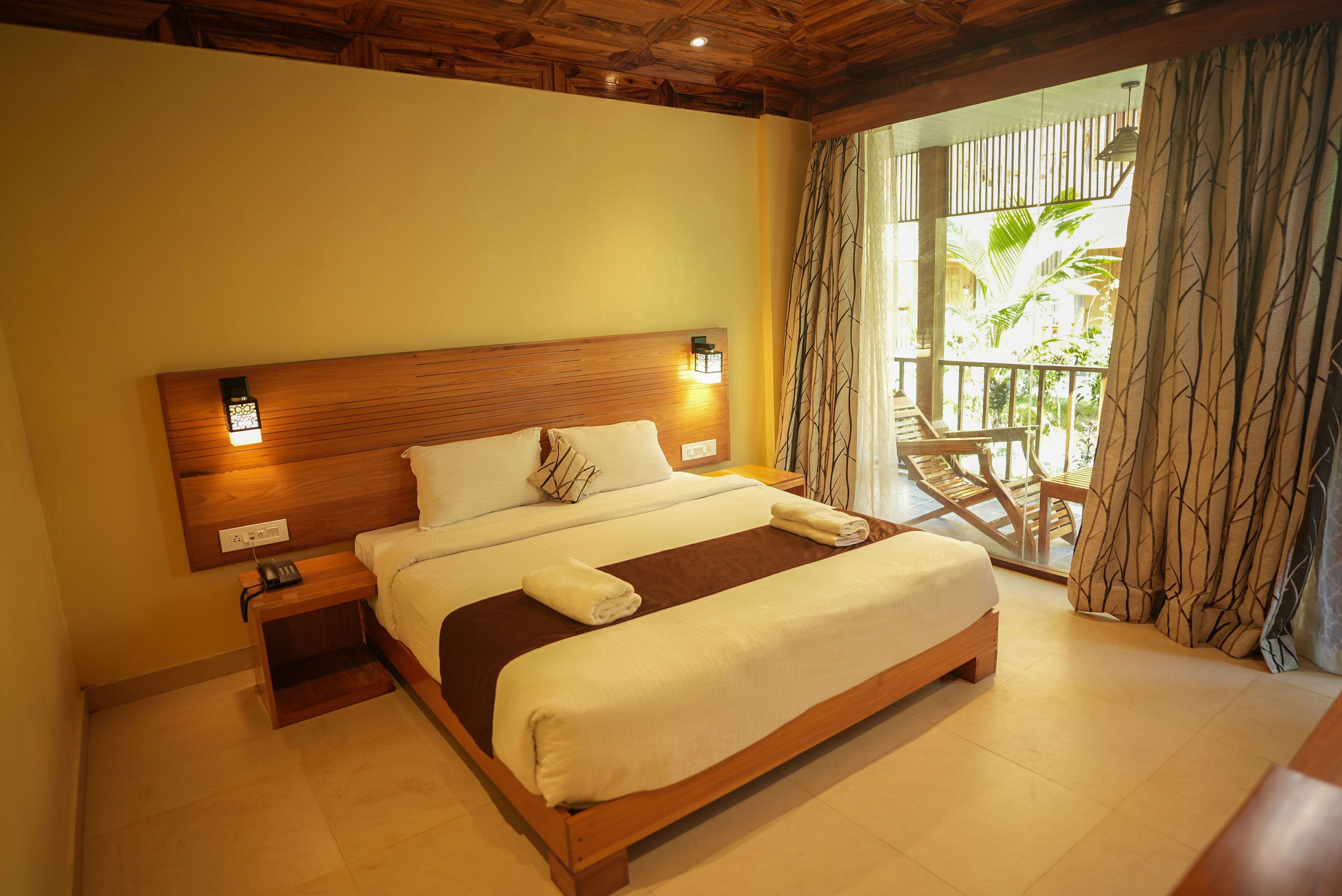 A neatly made bed with a wooden headboard, bedside lamps, and a view of a balcony with outdoor furniture.