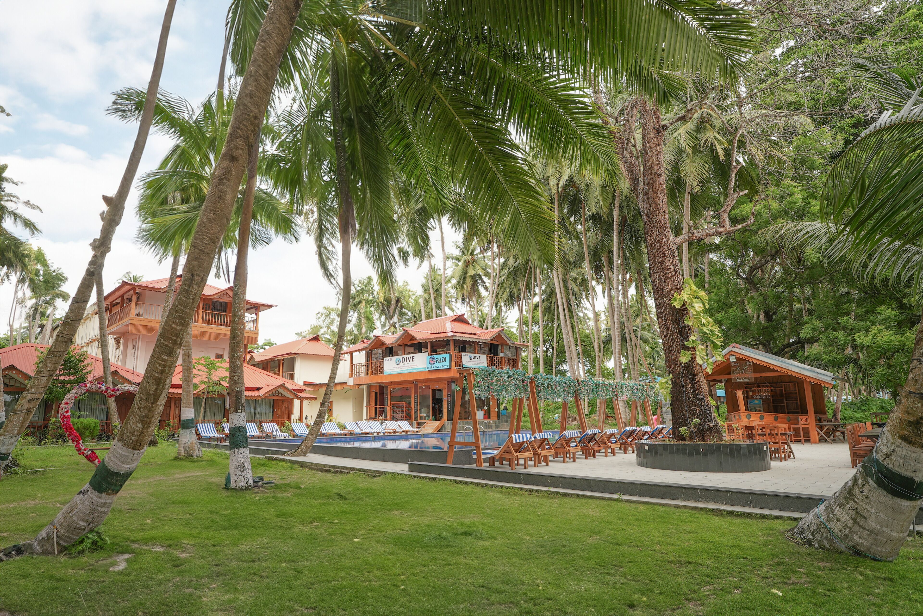 Un resort con varias edificaciones, un área de piscina con sillones de descanso y palmeras.
