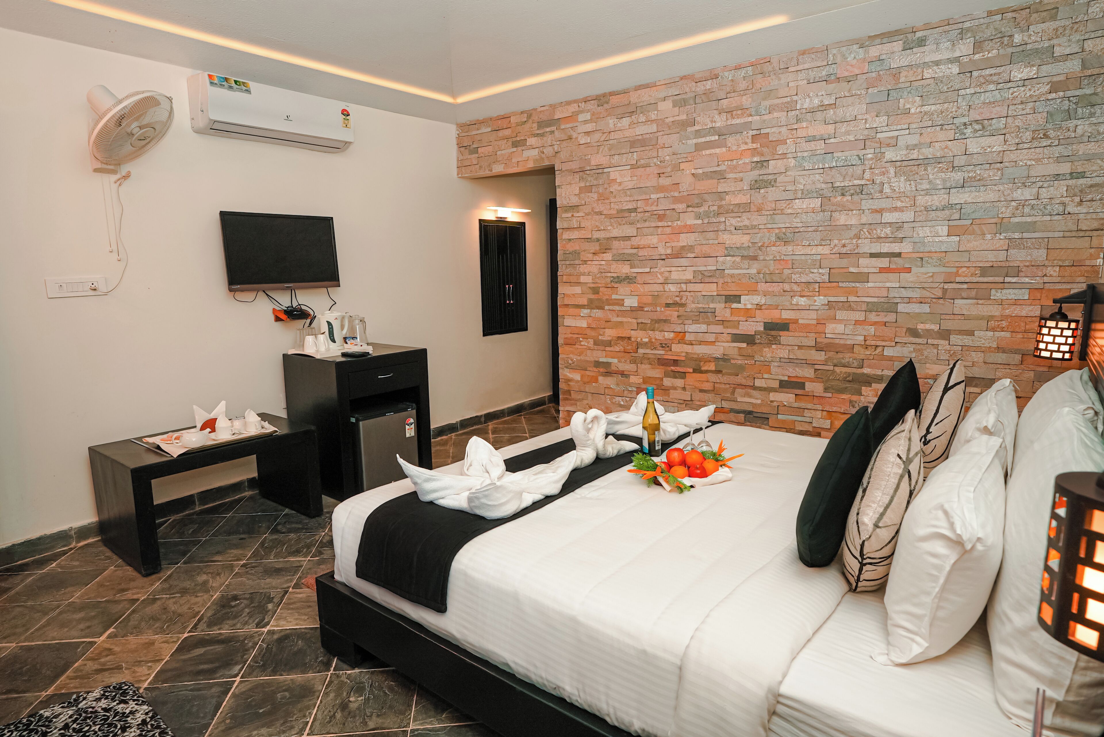 A hotel room with a brick-patterned wall, a bed with white bedding, a flat-screen TV, a desk with a fan, and a nightstand with toiletries.