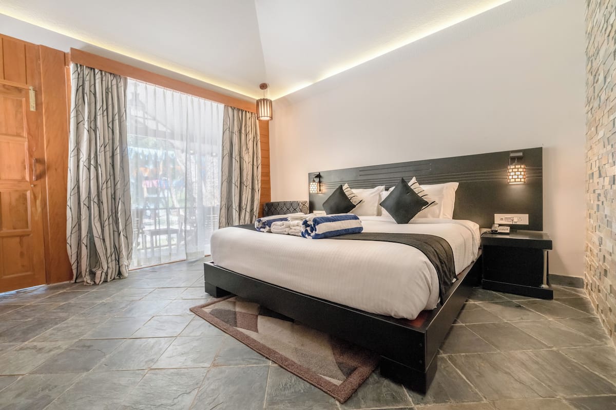 Quarto moderno com uma cama grande, uma mesinha de cabeceira, uma luminária na parede e vista pela janela com cortinas.