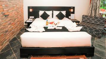 A neatly made bed with a bottle and swan-shaped towels, a nightstand with a phone, and a patterned sofa in a modern room with a brick wall and tiled floor.