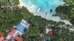 Vista aérea de un resort en la playa con piscina, palmeras y barcos en el agua.
