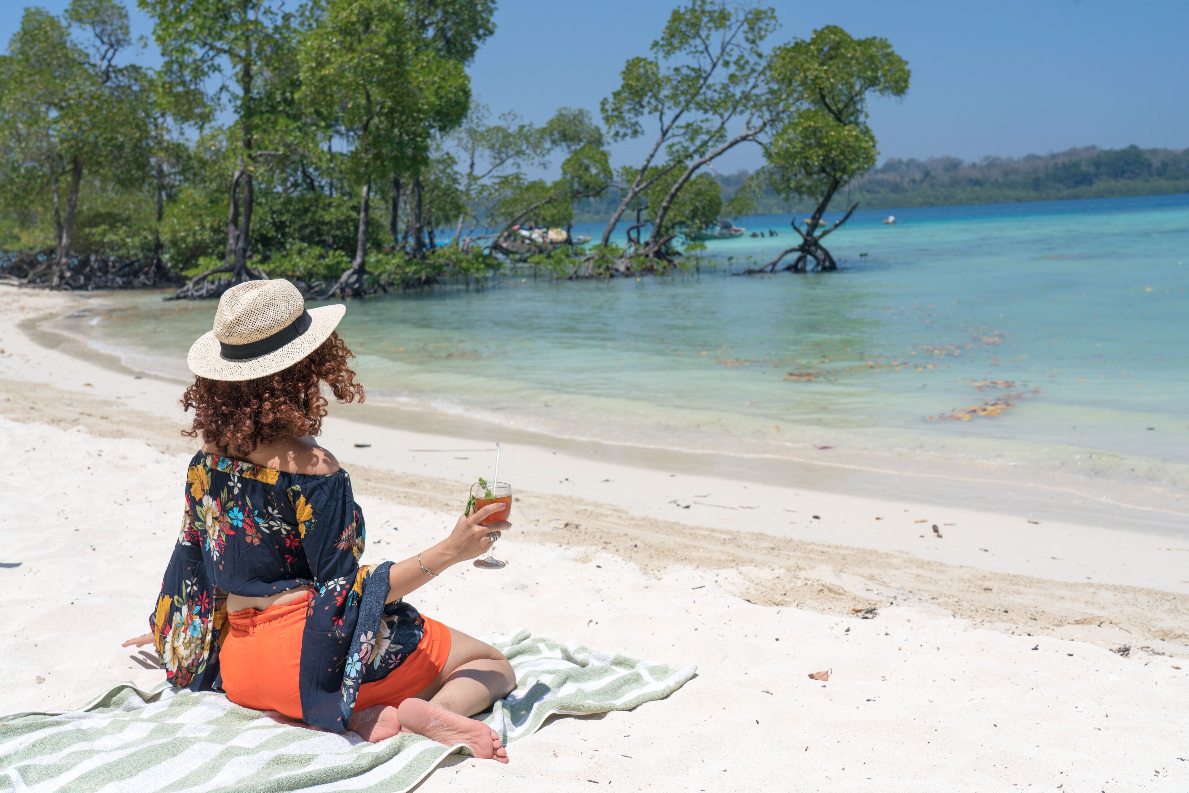 Une personne assise sur une serviette de plage, tenant une boisson, avec une vue dégagée sur l’océan et des arbres en arrière-plan.