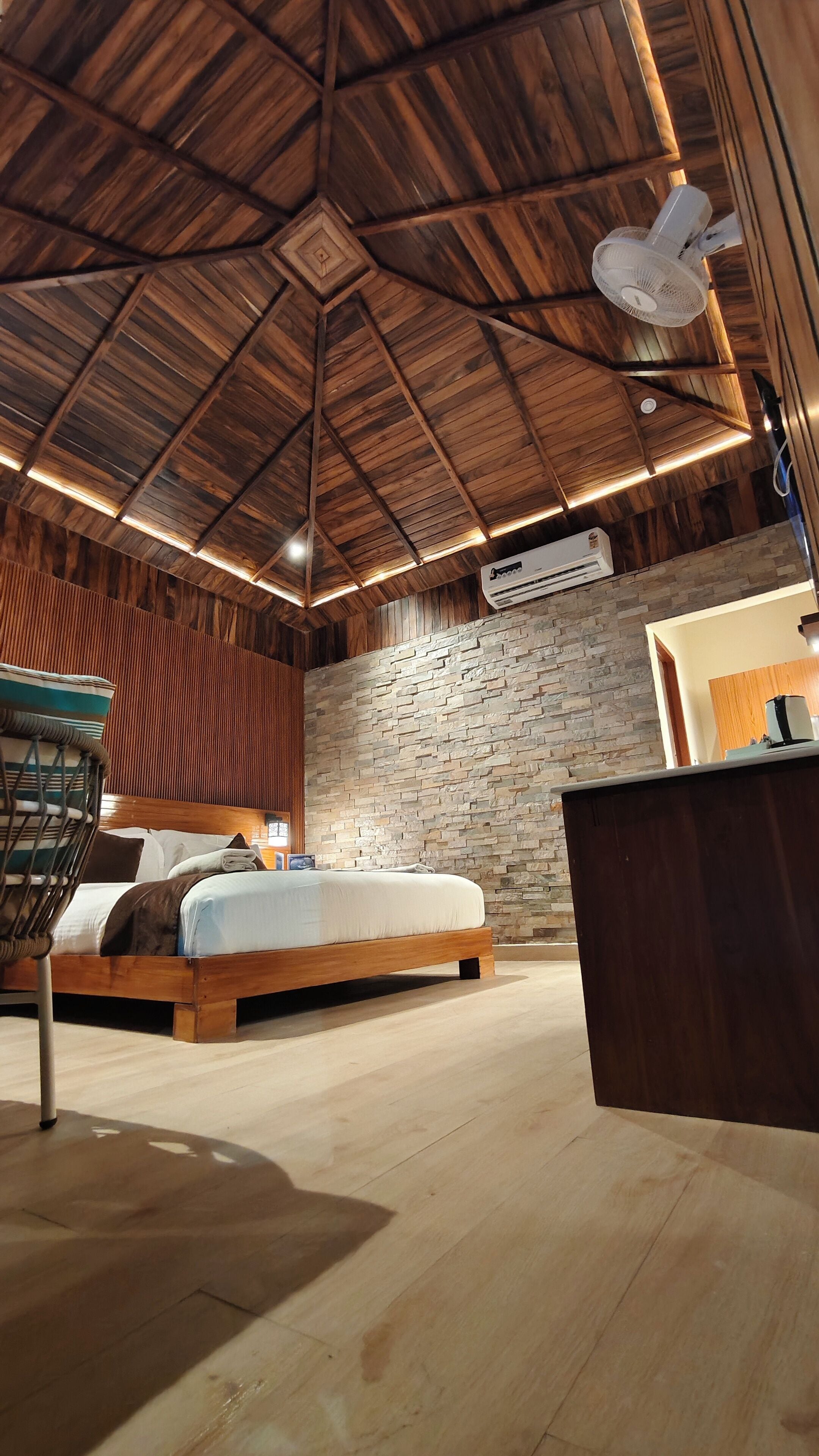 A spacious bedroom with a wooden ceiling, a stone wall, a bed, a chair, and a desk with a lamp.