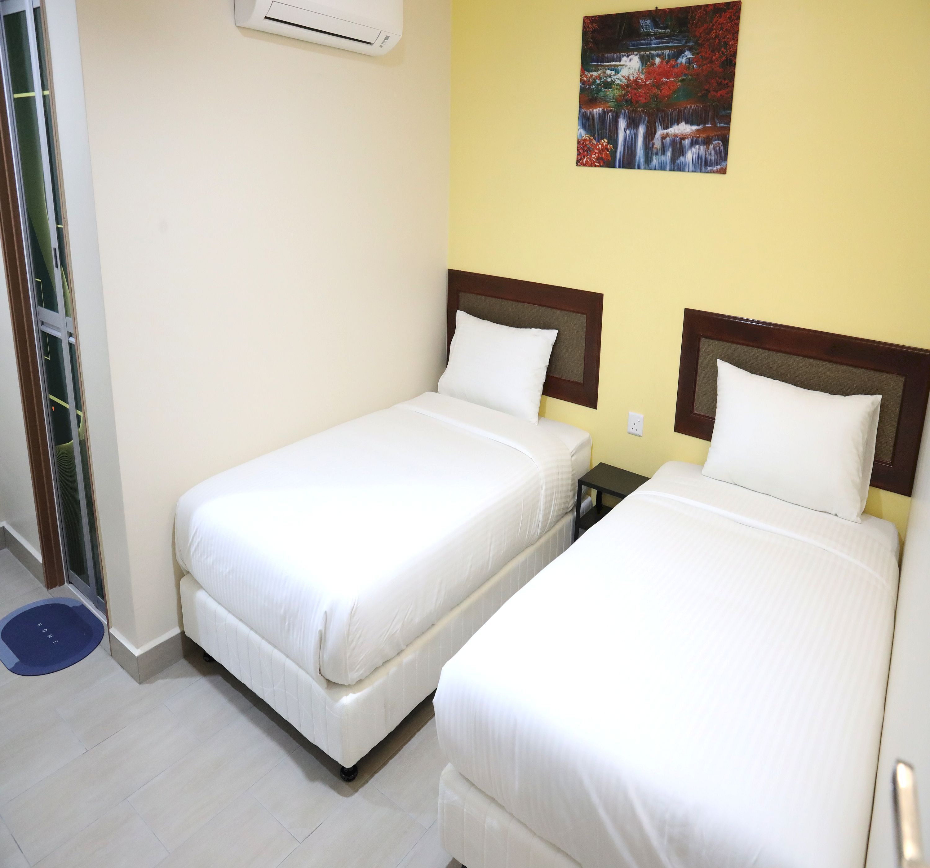 Habitación de hotel con dos camas individuales, una mesita de noche pequeña, aire acondicionado de pared y un cuadro de una cascada.