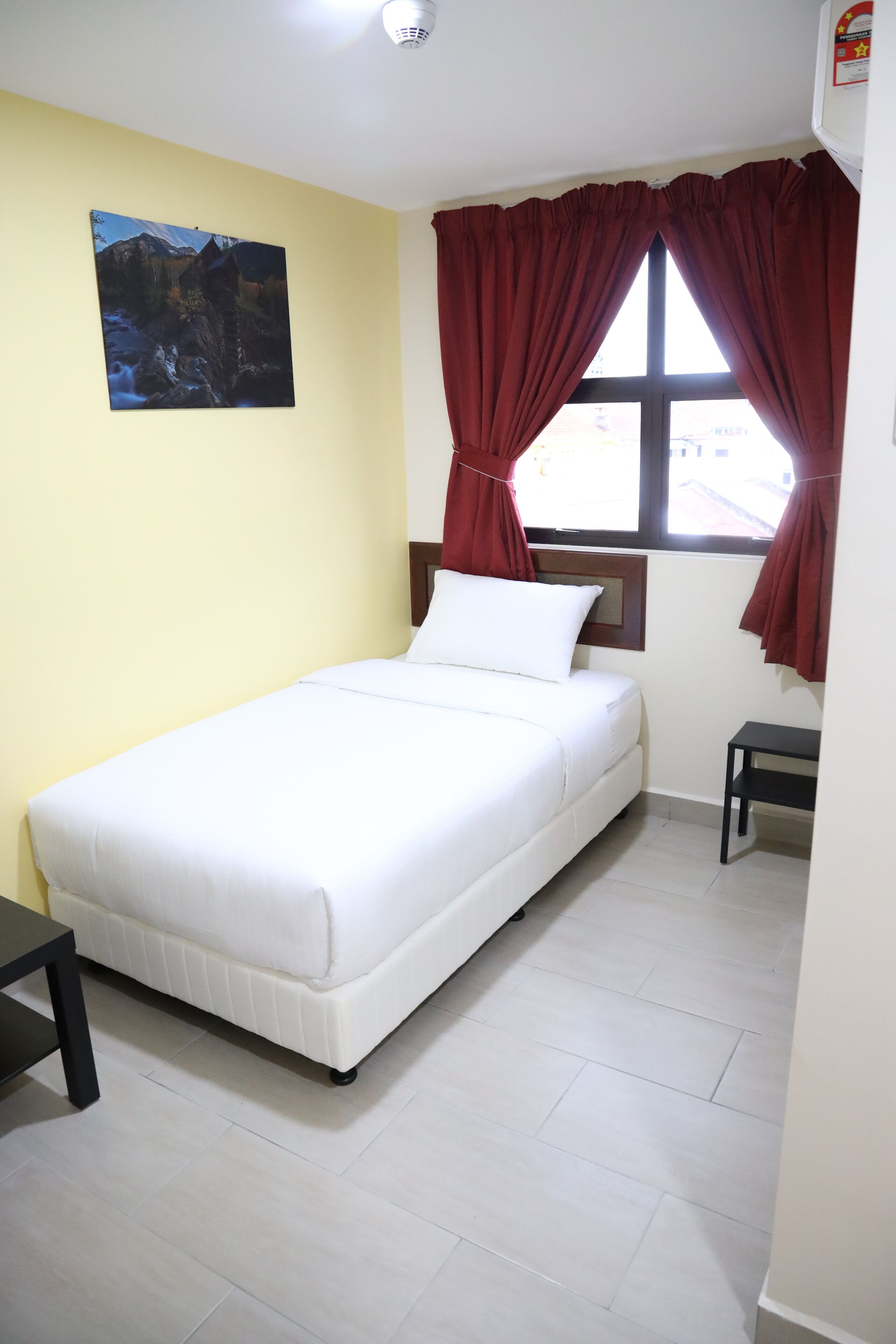 A single-bed room with a white mattress, a small bedside table, a nightstand, a window with red curtains, and a painting on the wall.