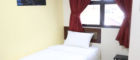 A single-bed room with a white mattress, a small bedside table, a nightstand, a window with red curtains, and a painting on the wall.