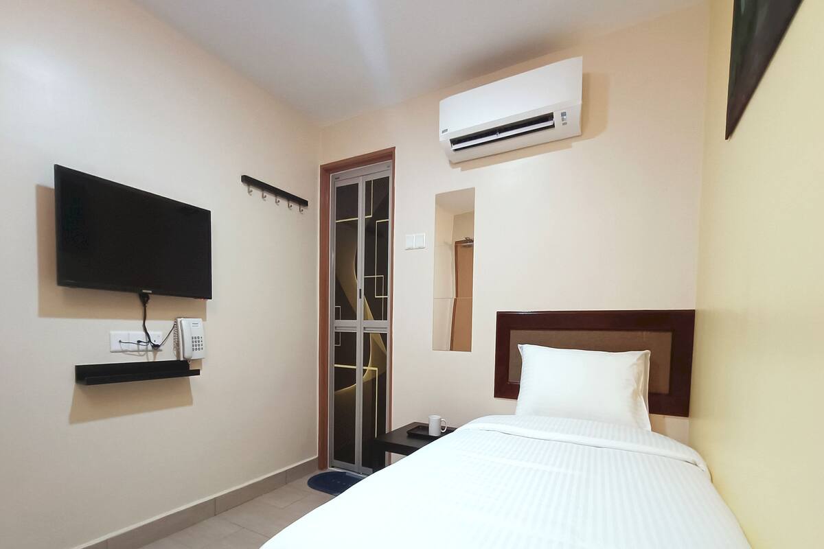 A hotel room with a bed, a television mounted on the wall, an air conditioning unit, a mirror, and a small table with a cup.