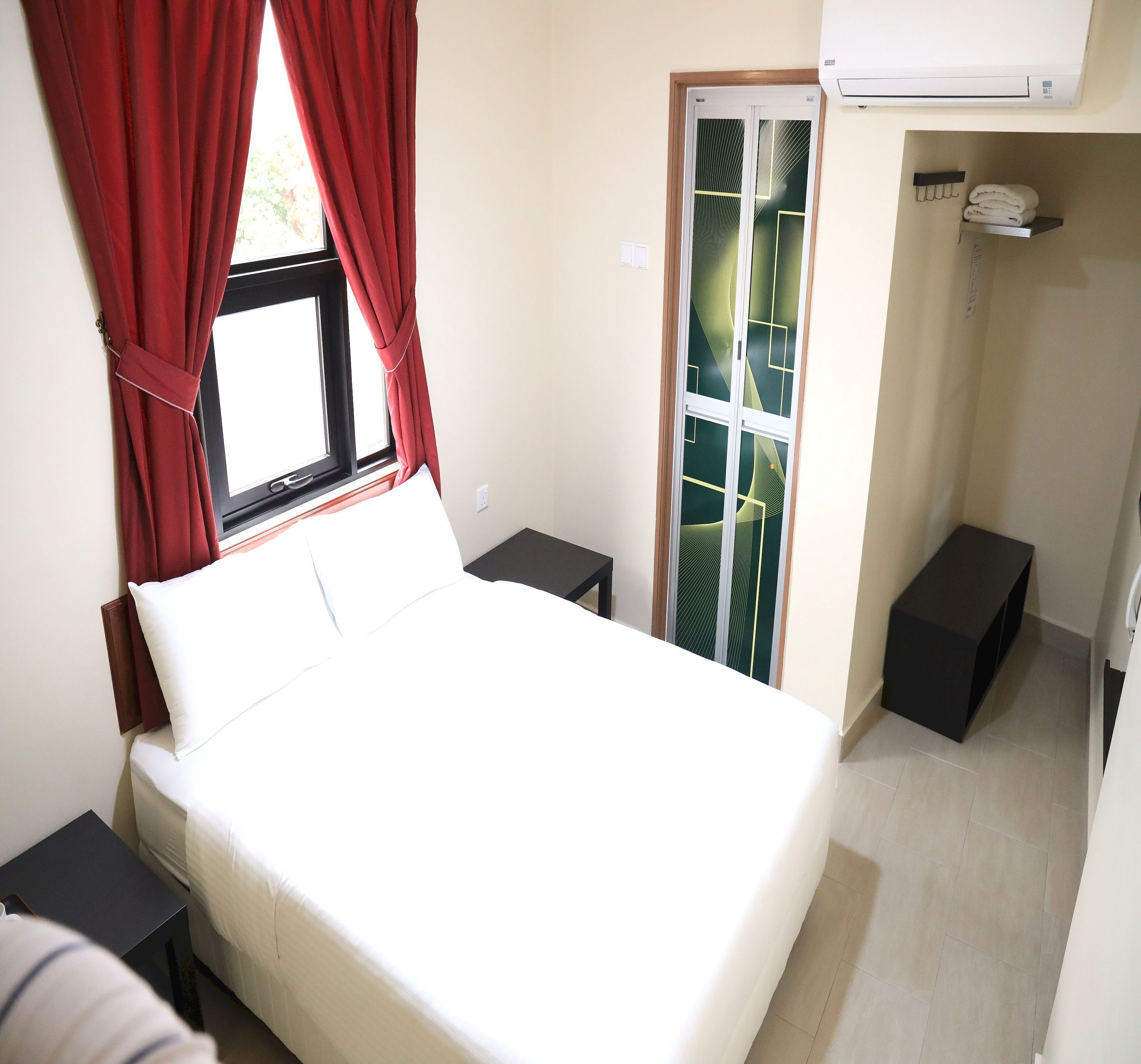 A bedroom with a bed, a window with red curtains, a sliding glass door, a small bedside table, and a black storage box.