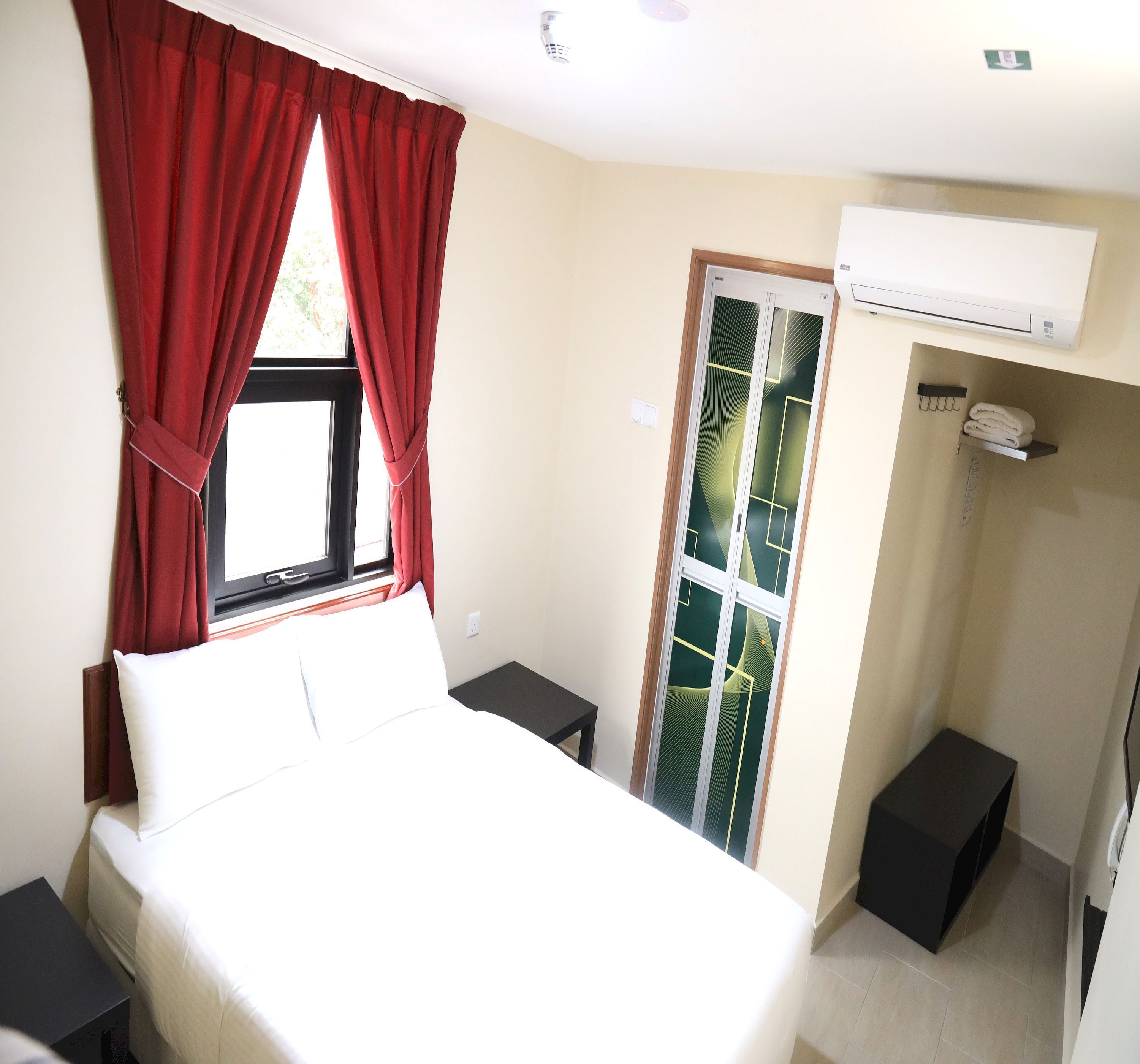 Une chambre d’hôtel avec un lit, une fenêtre avec des rideaux rouges, une armoire et un système de climatisation intégré.