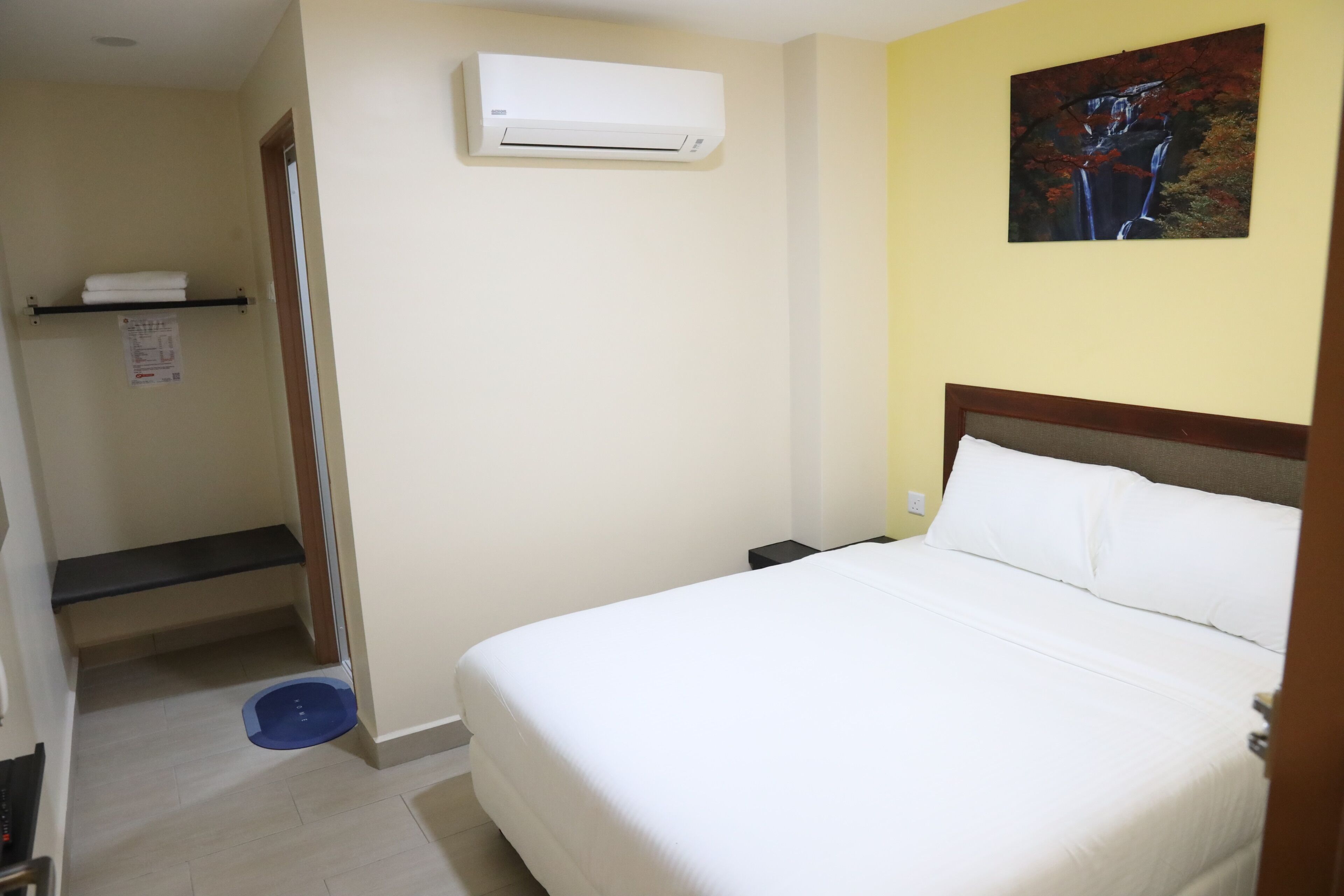 A hotel room with a bed, an air conditioner, a painting on the wall, and a shelf with towels.