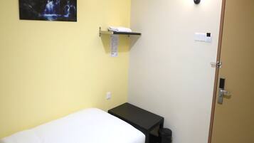 A small, simple room with a bed, a small table, a trash bin, a door, and a wall-mounted shelf with a towel.