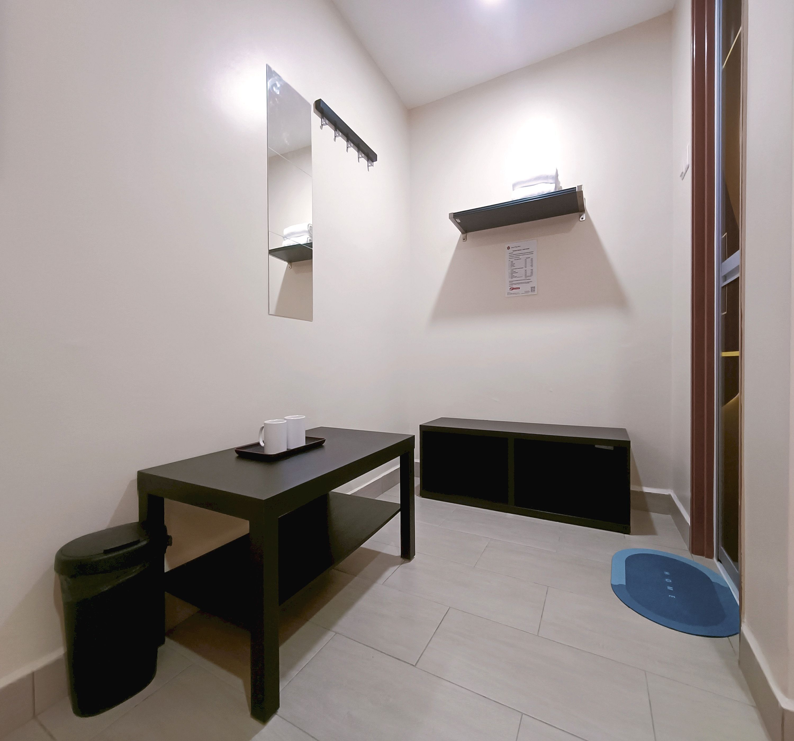 Une petite pièce propre, avec un bureau, une poubelle, un miroir et un tapis bleu sur le sol.