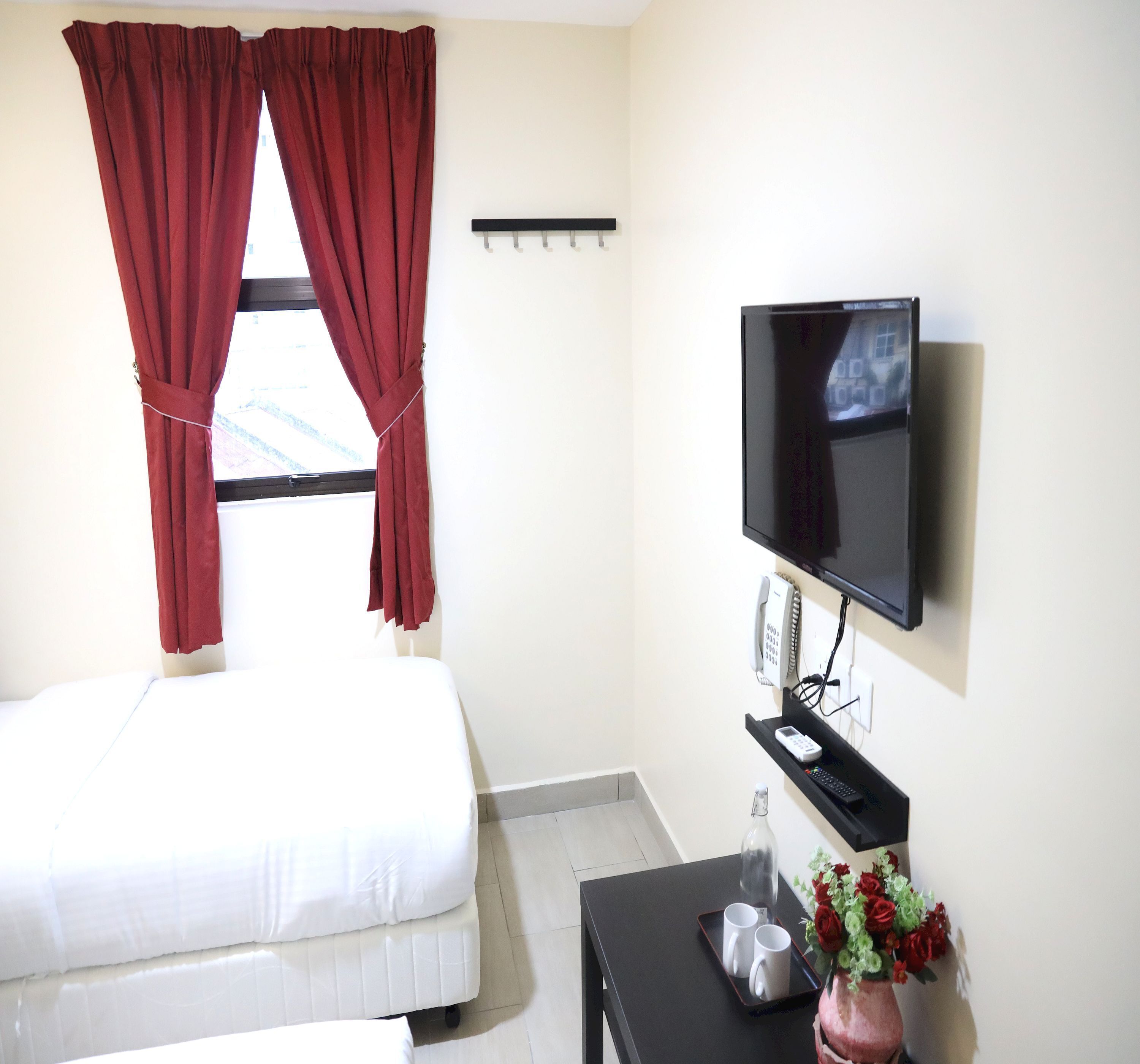 Ein Hotelzimmer mit einem Bett, einem an der Wand montierten Fernseher, einem Nachttisch mit Telefon und einer Blumenvase.